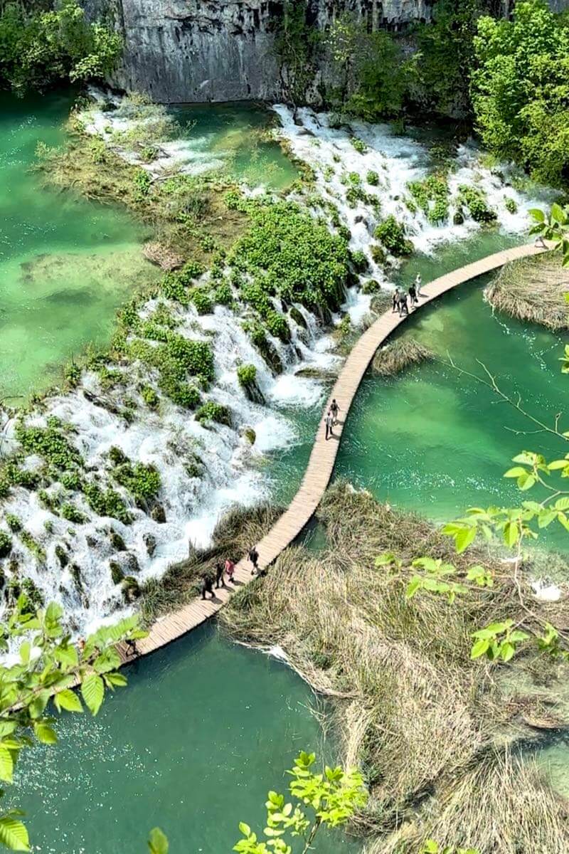 Aerial view of the most famous and picturesque part of Lower Lakes of Plitvice Lakes National Park, Croatia. People walk on the windy boardwalk in the middle of lakes next to low and layered waterfalls weaving through greens.